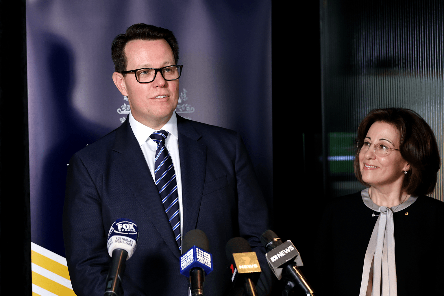 Kieren Perkins speaks in front of broadcast microphones while Josephine Sukkar watches.