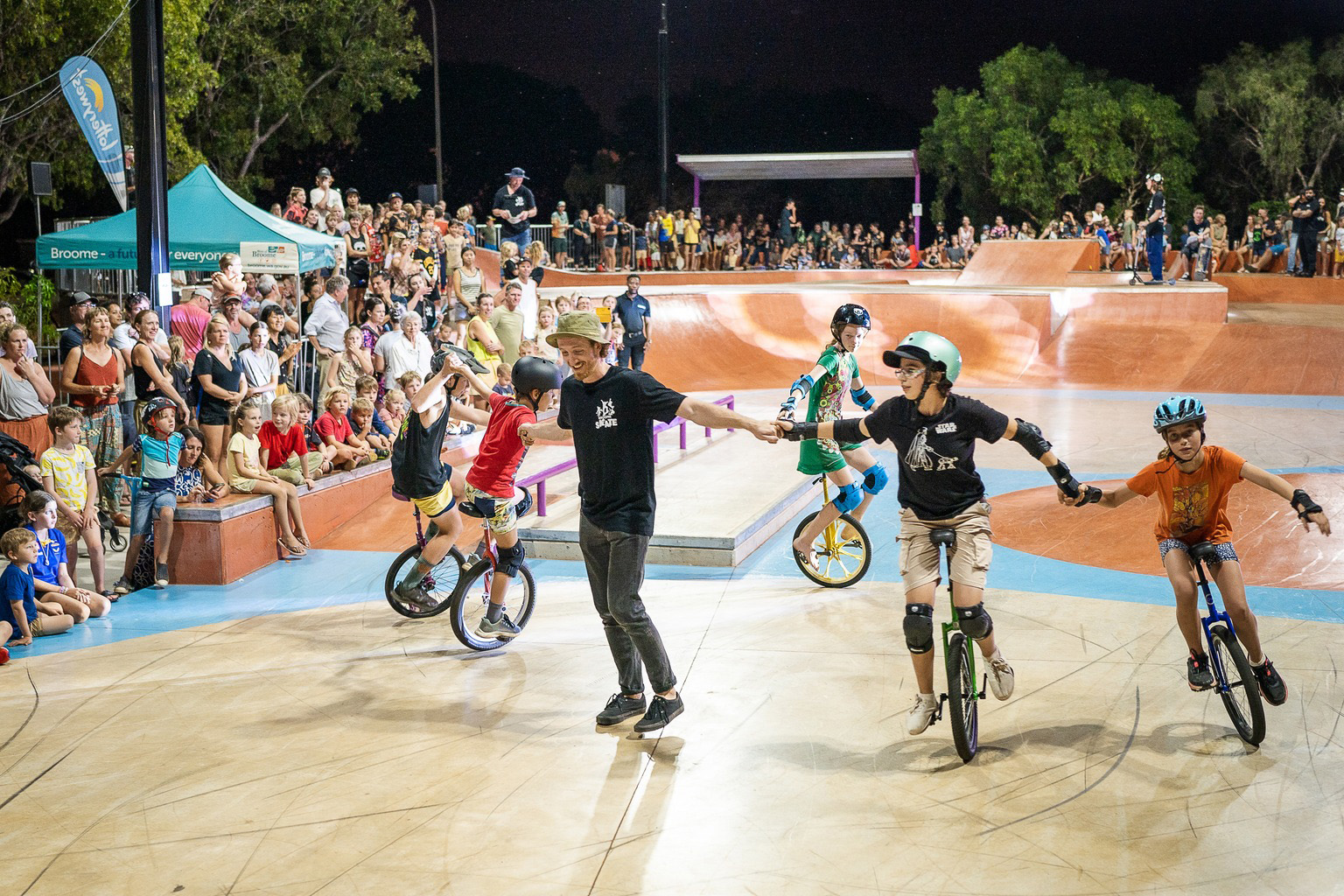 A instructor leads several unicyclists through a demonstration at Broome Skate Park in front of a large crowd.