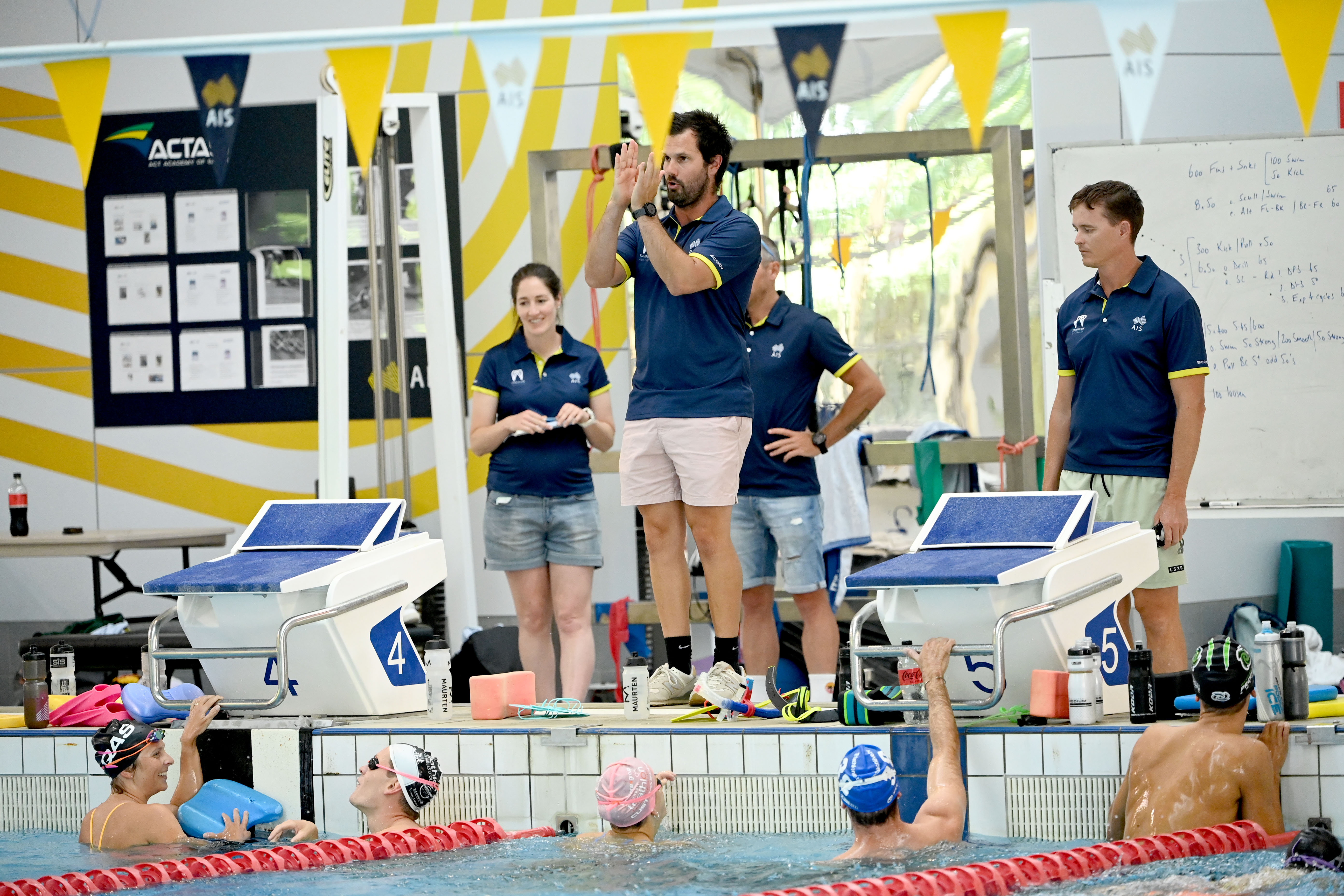 A coach stands on the edge of the pool coaching athletes in the pool. 