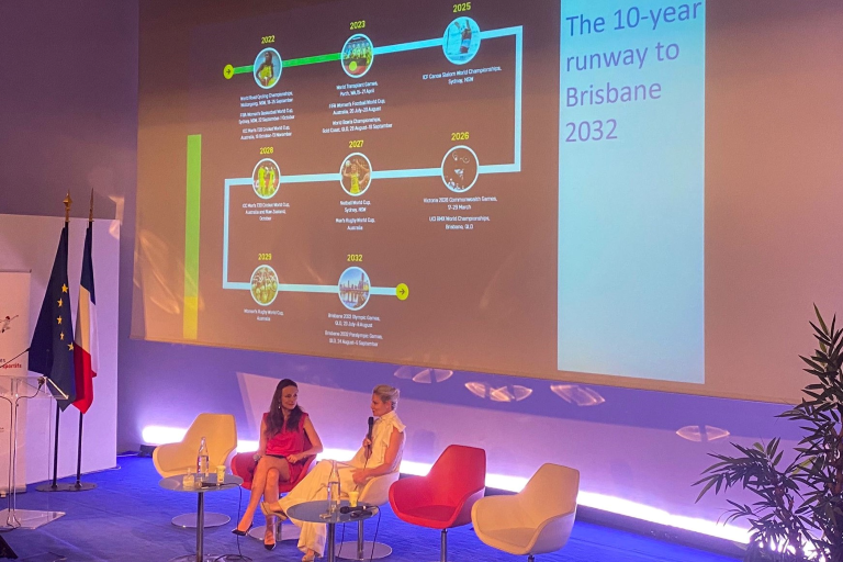 Two women sit on stage in discussion with a presentation screen behind them