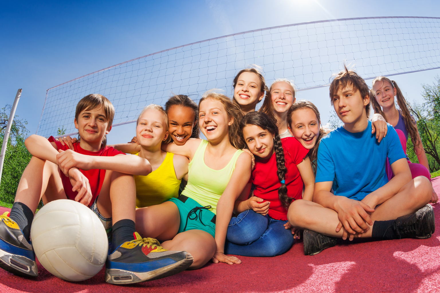 A group of teenage children sitting huddled together smiling with a volleyball, in front of a net.