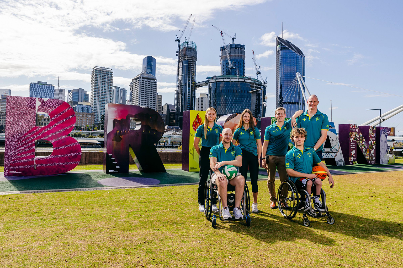 A group of paralympic athletes gathered in front of large letters spelling Brisbane, with the Brisbane skyline in the background.
