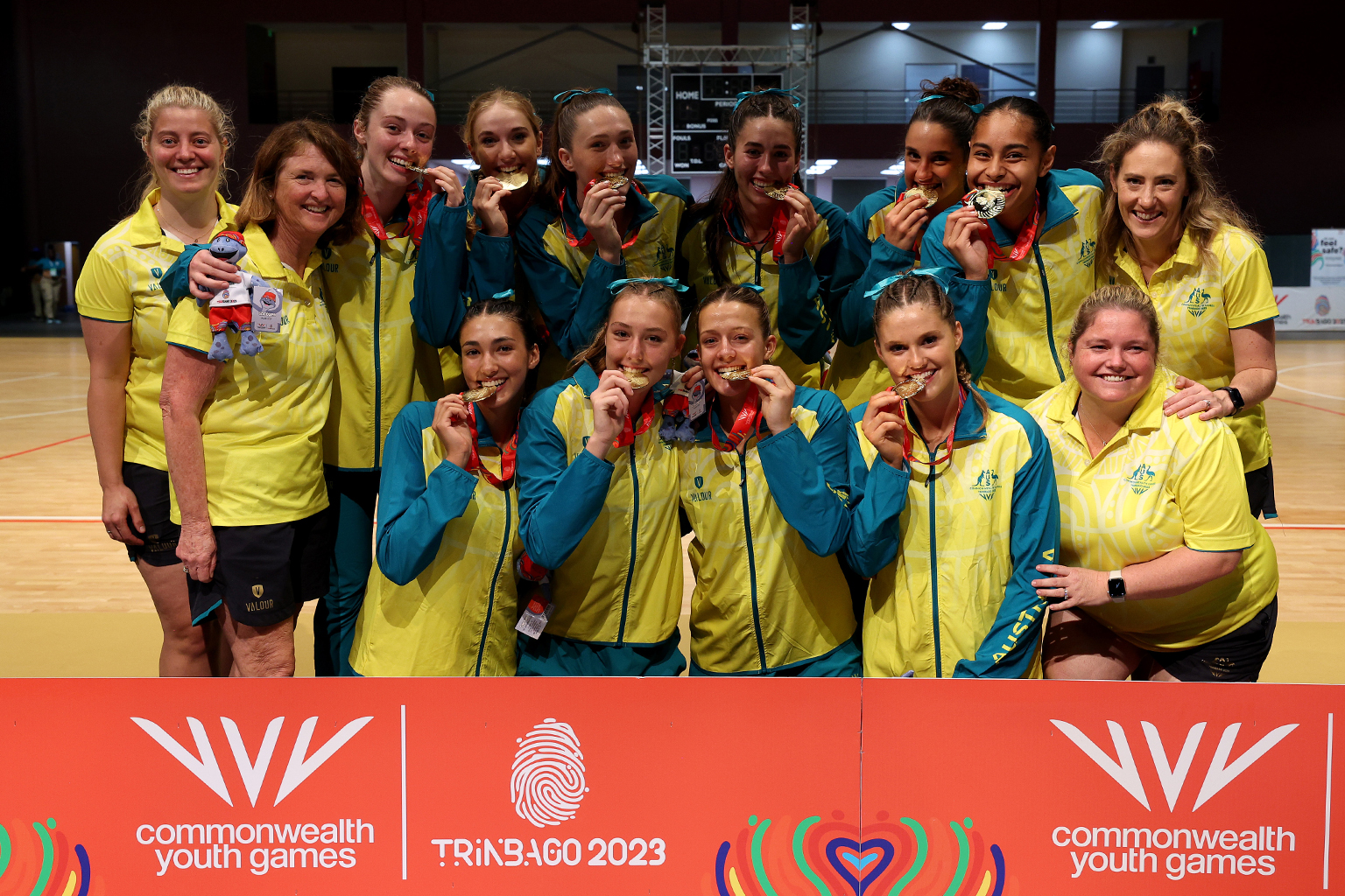 A team of athletes huddled together on a netball court, behind Commonwealth Youth Games Trinbago 2023 signage, playfully biting on their gold medals.