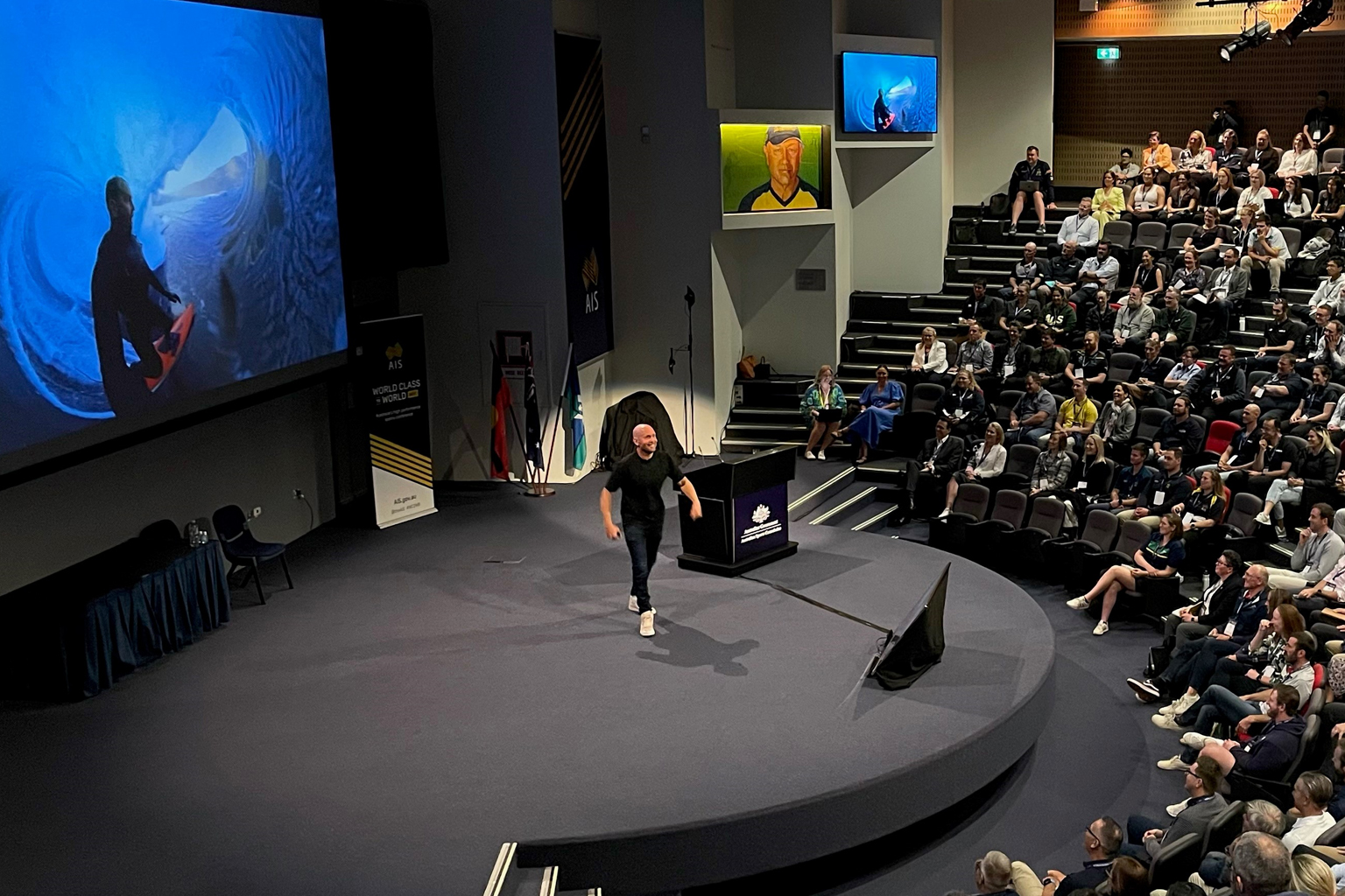 A man stands on stage, speaking, with a screen showing an image of a surfer