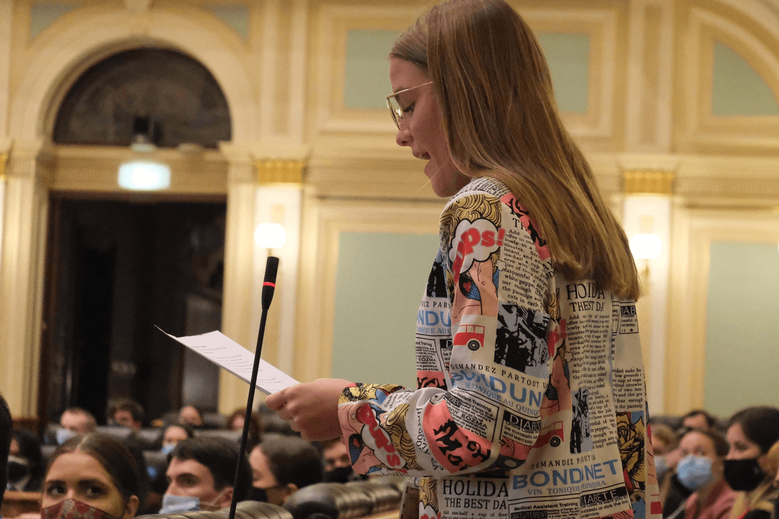 Side image of Imogen Ruyg standing at a lectern with other people seated around her, wearing face masks.