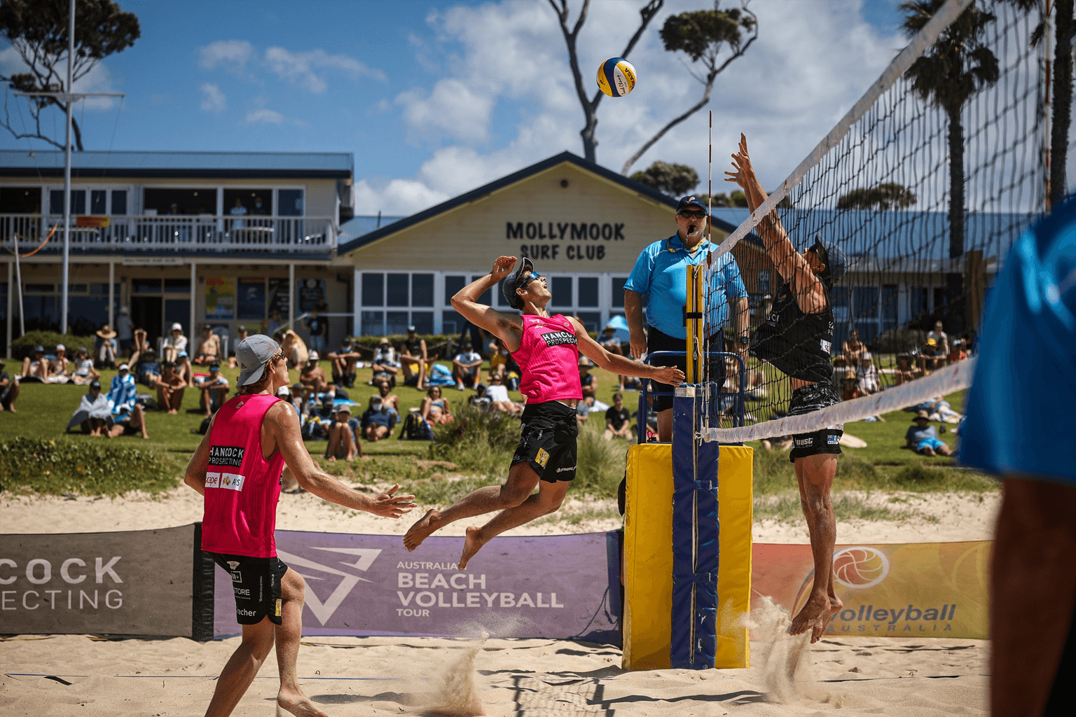 Men play in a beach volleyball match in front of Mollymook Surf Club and spectators sitting on a lawn.