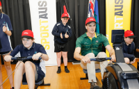 A Melba Copeland Secondary School student and Olympic rower Alex Purnell use rowing machines in front of SportAUS banners and Australian, Aboriginal and Torres Strait Island flags.