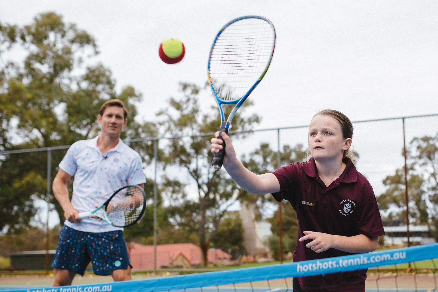 A student hits a tennis ball over the net with Matt Ebden watching in the background.