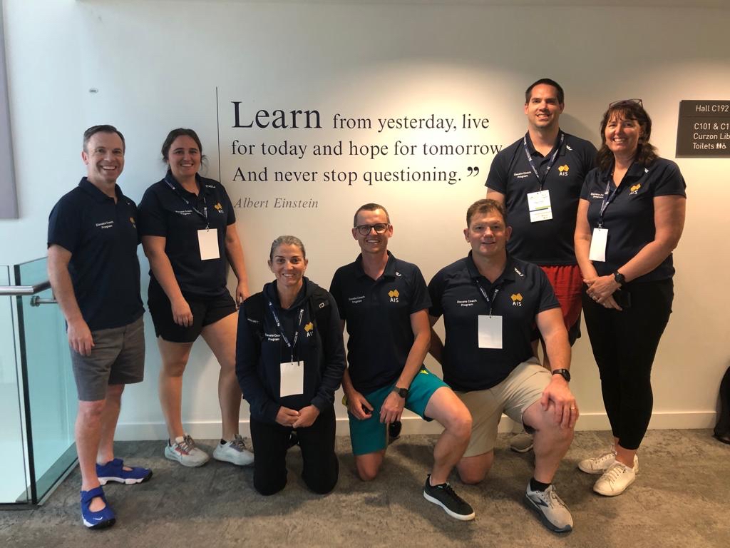 Six coaches stand in front of a sign which reads 'Learn'. 