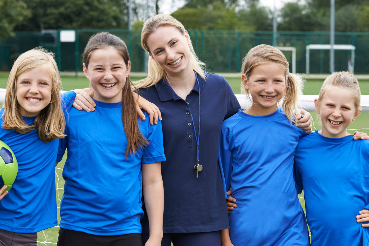 Four girls in blue team uniforms stand arm in arm with a woman wearing a whistle around her neck. One of the girls holds a soccer ball.