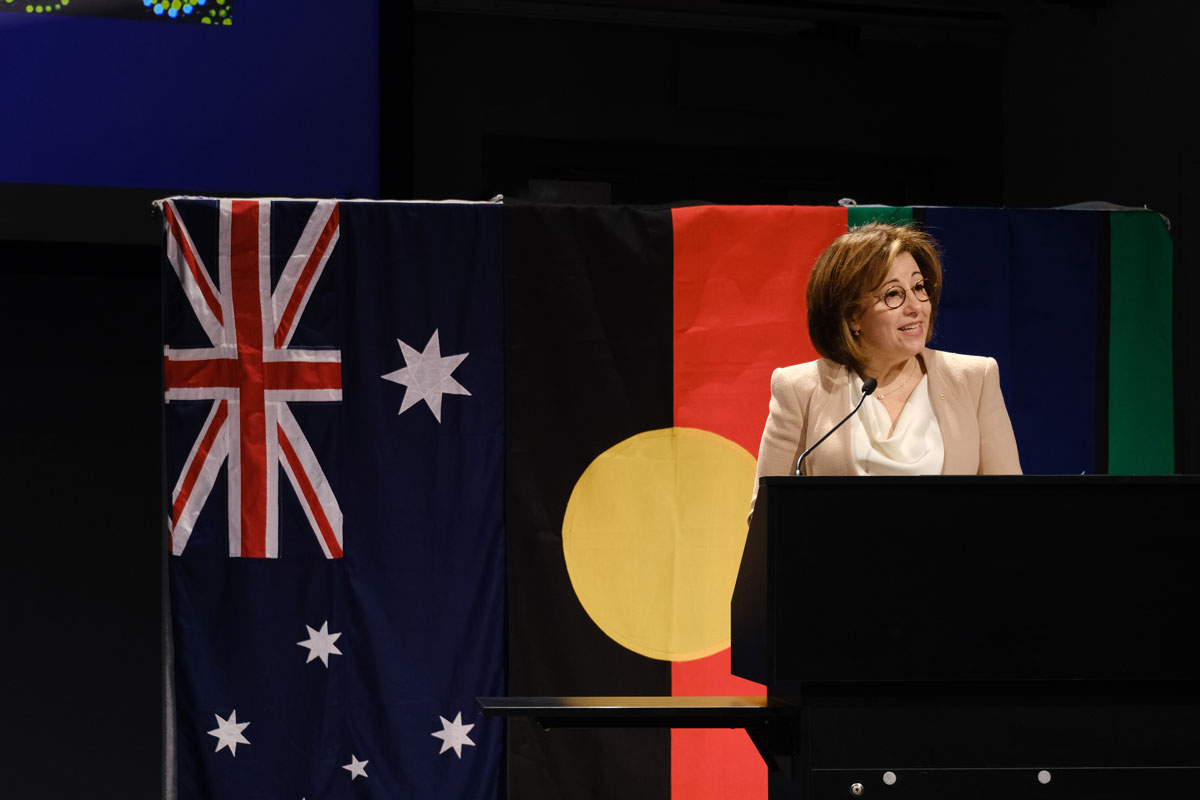 Josephine Sukkar stands at a lectern in front of Australian, Aboriginal and Torres Strait Island flags