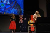 Wiradjuri Echoes performer Jakida Smith stands on stage wearing traditional paint while Duncan Smith stands behind a lectern wearing traditional Wiradjuri clothing and paint. Australian, Aboriginal and Torres Strait Island flags and an Aboriginal artwork are behind them.