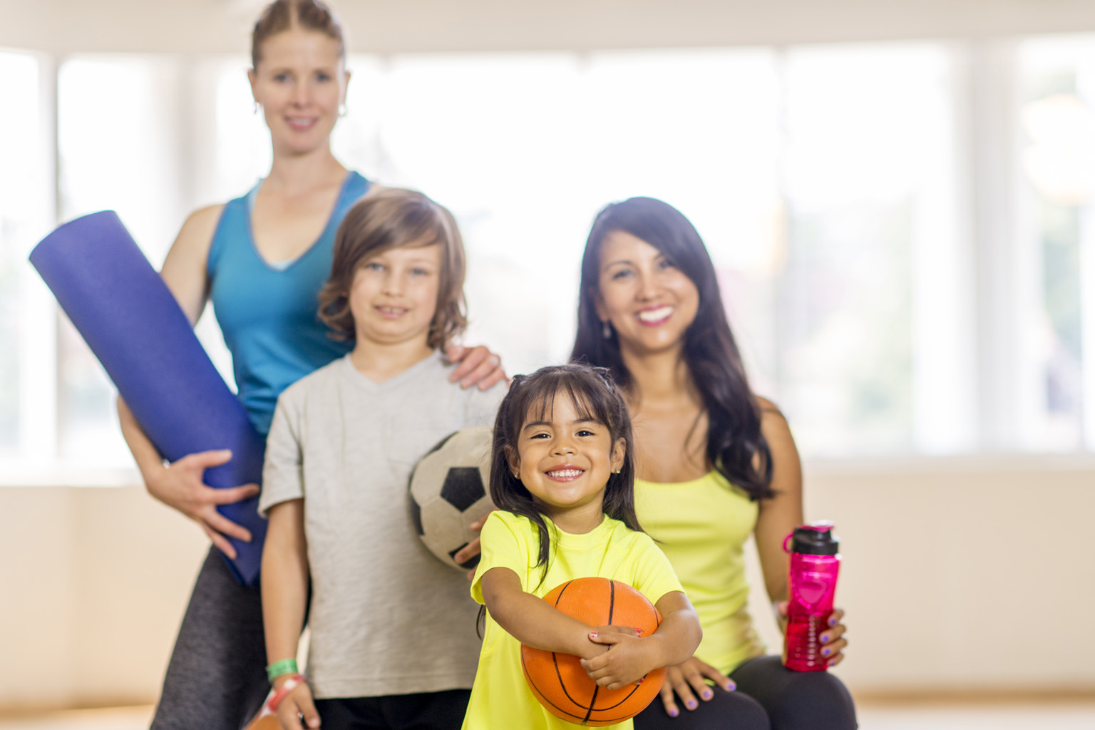 A woman holding a yoga mat, a boy with a soccer ball, a young girl with a basketball and a woman holding a sports drink bottle