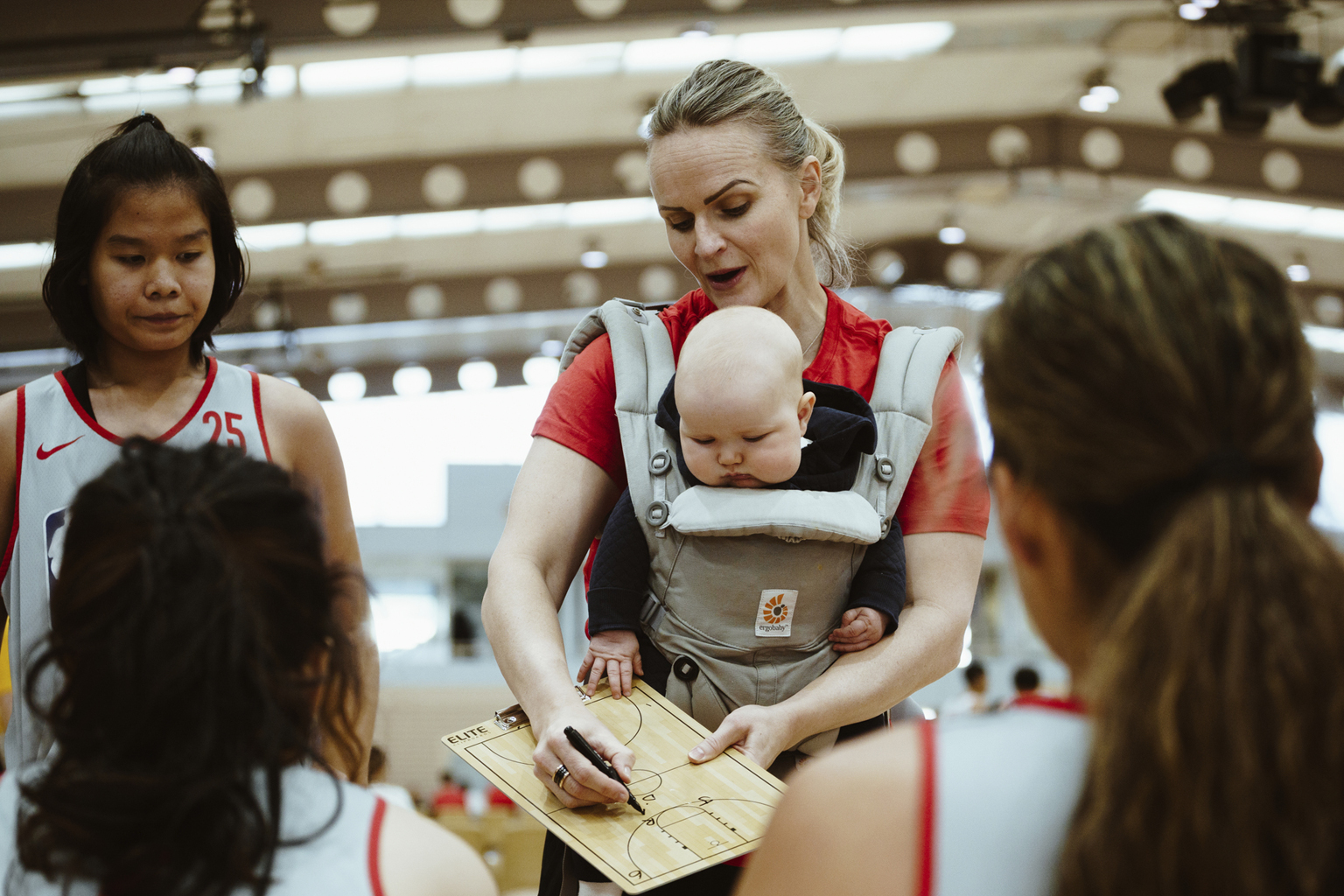 A woman coaches a basketball team, while holding a baby in a carrier on her front.. 