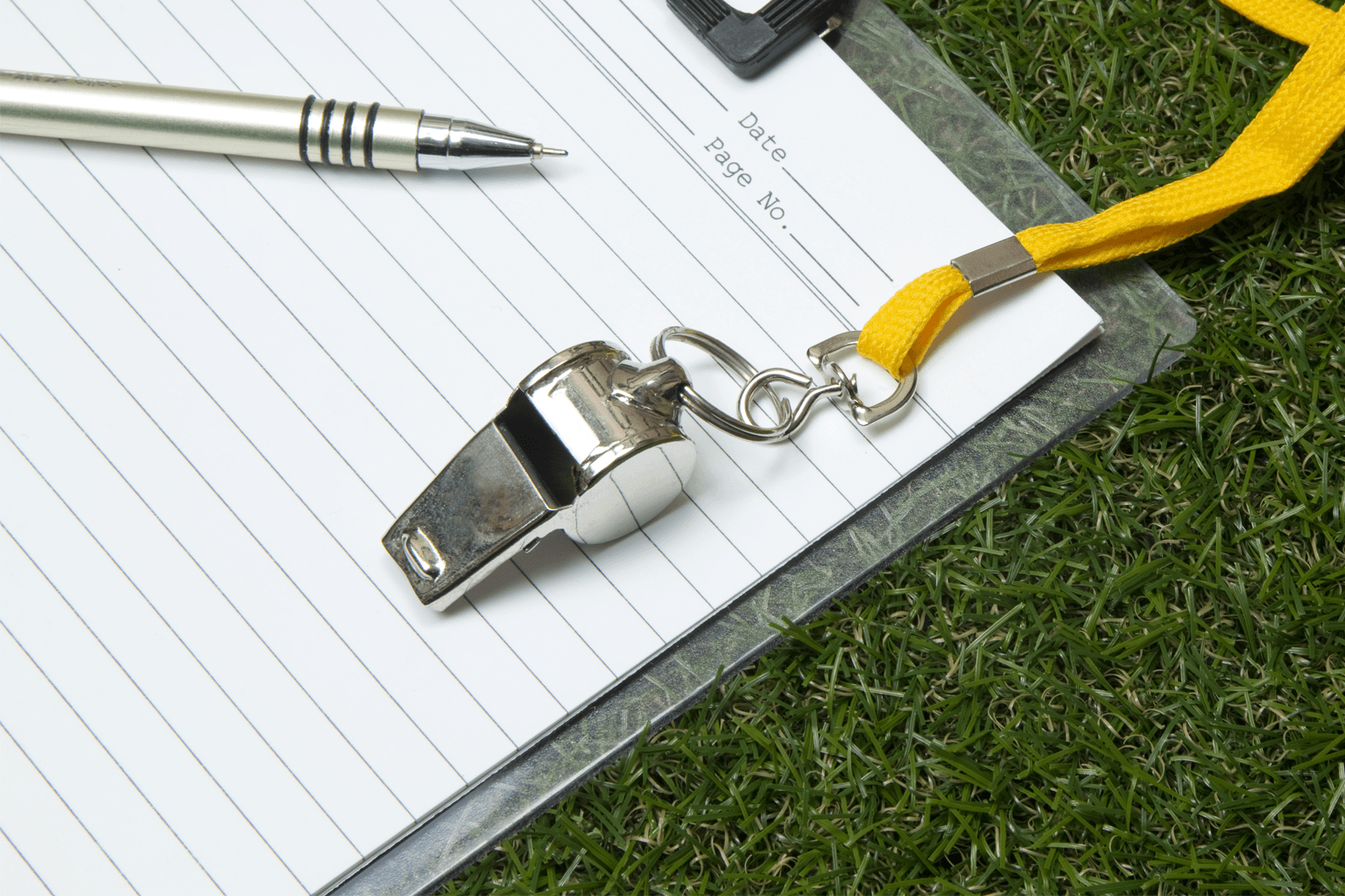 Notepad and pen sitting on grass with a referee's whistle.