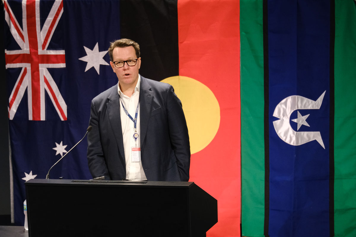 Kieren Perkins stands at a lectern in front of Australian, Aboriginal and Torres Strait Island flags