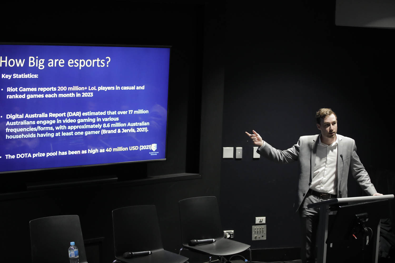 A man in a grey suit stands at a lectern, pointing to a screen with the title 'how big are esports? Key statistics: Riot Games reports 200m+ players in casual and ranked games each month in 2023. Digital Australia Report estimated that over 17m Australians engage in video gaming in various forms, approx 8.6m Aust housholds have at least 1 gamer/ the DOTA prize pool has been as high as $40m US.