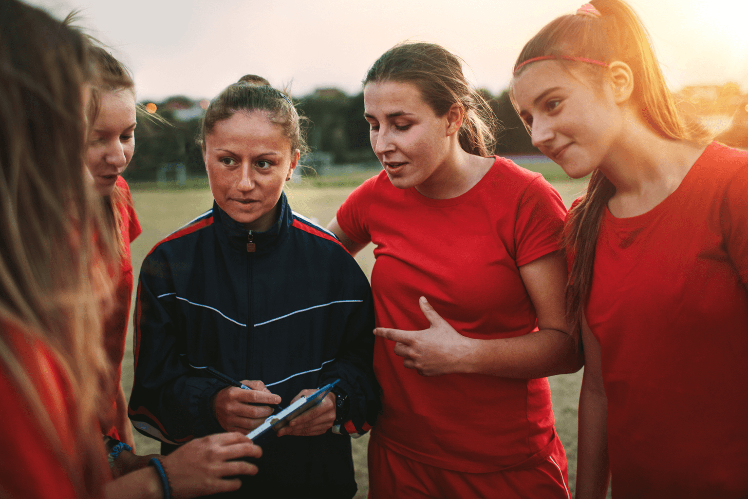 A woman holding a clipboard speaks to players in a women's sports team.