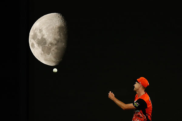 Image of a moon, a white cricket ball and Jason Behrendorff of the Perth Scorchers fielding against a black night sky.