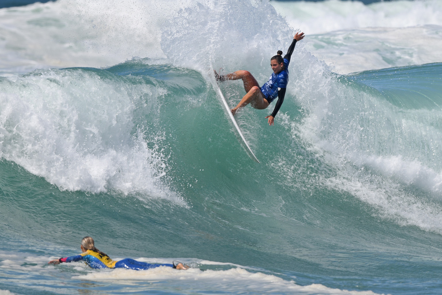 A woman surfs high on a wave while another woman paddles on her surfboard in the foreground