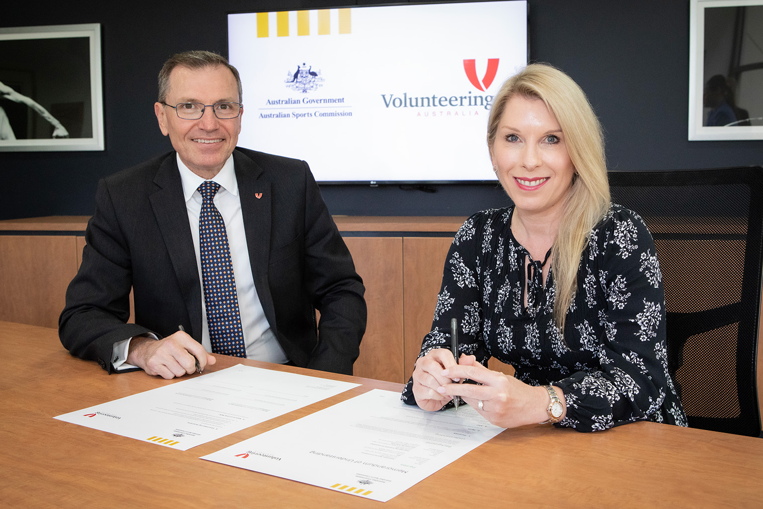Mark Pearce and Brooke De Landre at a desk signing papers. A screen in the background displays Australian Sports Commission crest and Volunteering Australia logo.