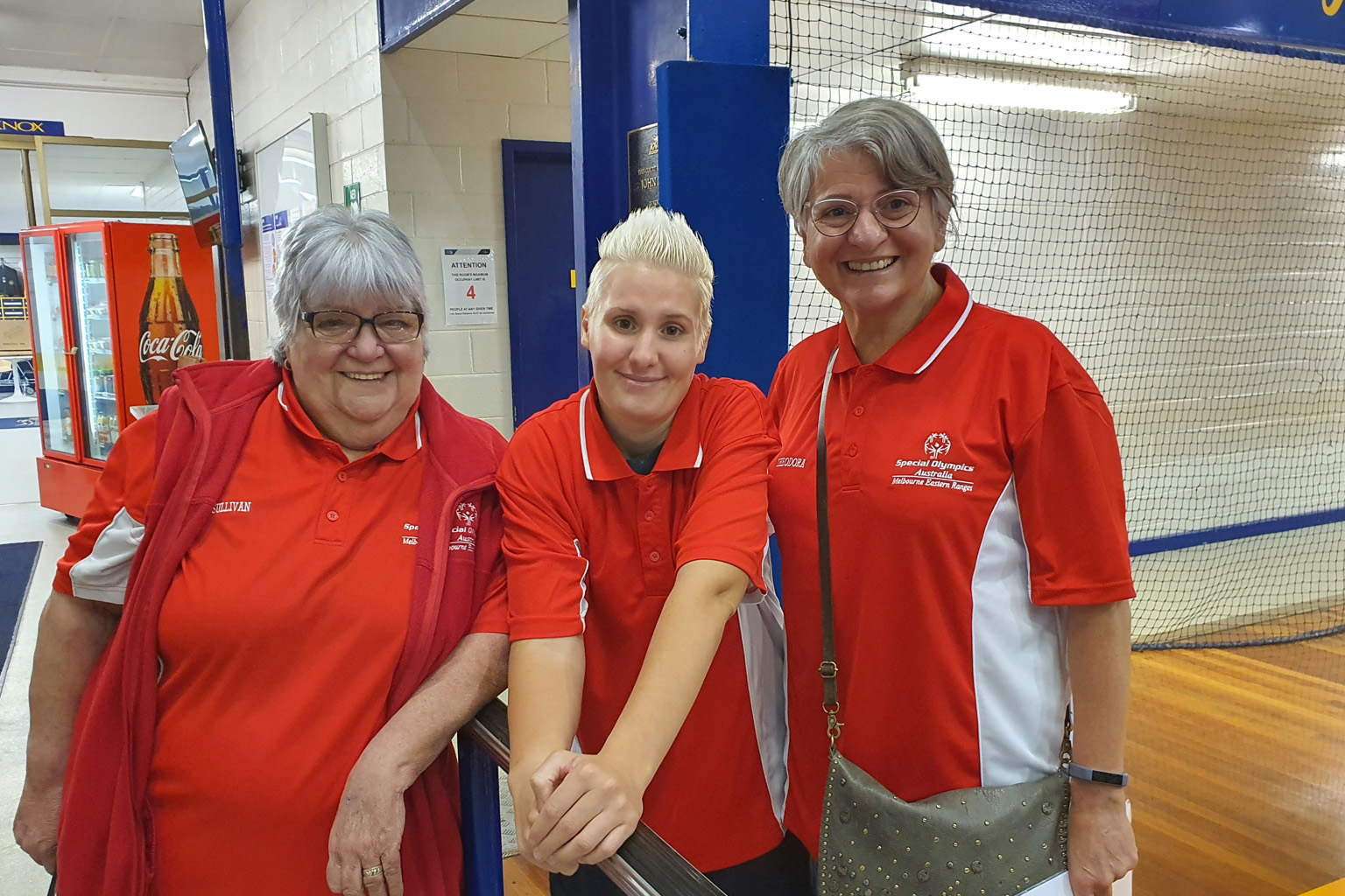 Three women stand together in an indoor sports facility wearing Special Olympics Australia Melbourne Eastern Rangers T-shirts
