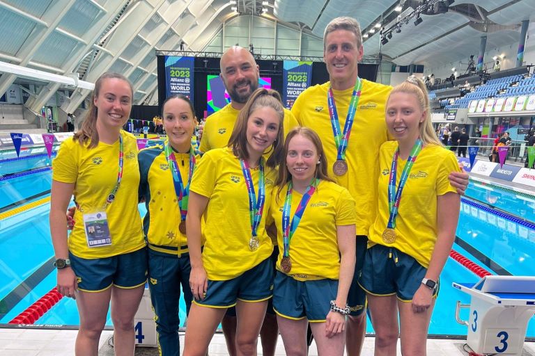 Casey Atkins stands in front of a swimming pool with a group of athletes at the 2023 Para World Swimming Championships.