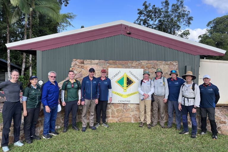 A group of coaches stand in front of a shed with a sign that reads 'Combat Training Centre.'