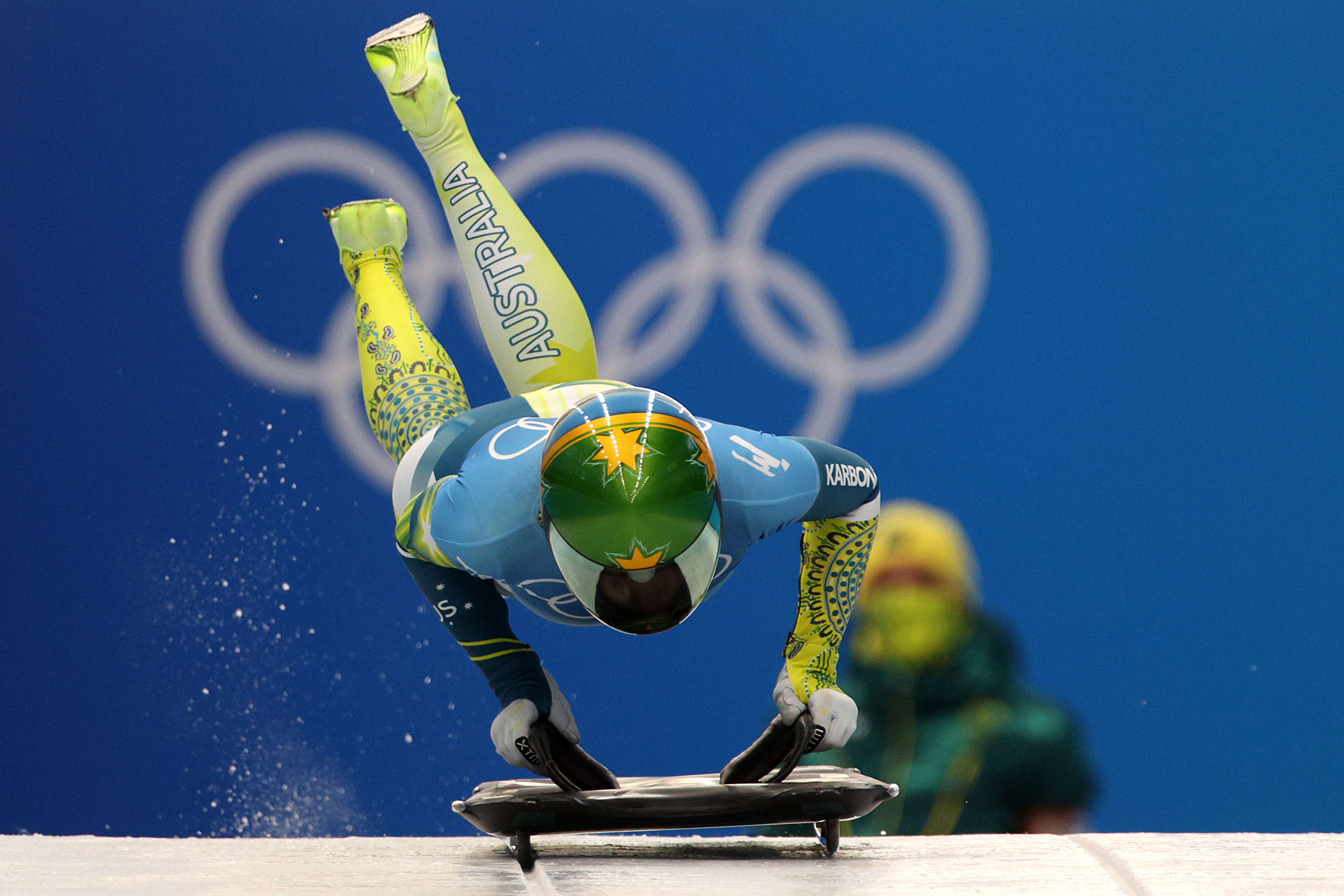 Image shows a skeleton racer jumping into their sled. The racer is wearing a green and gold skinsuit. 