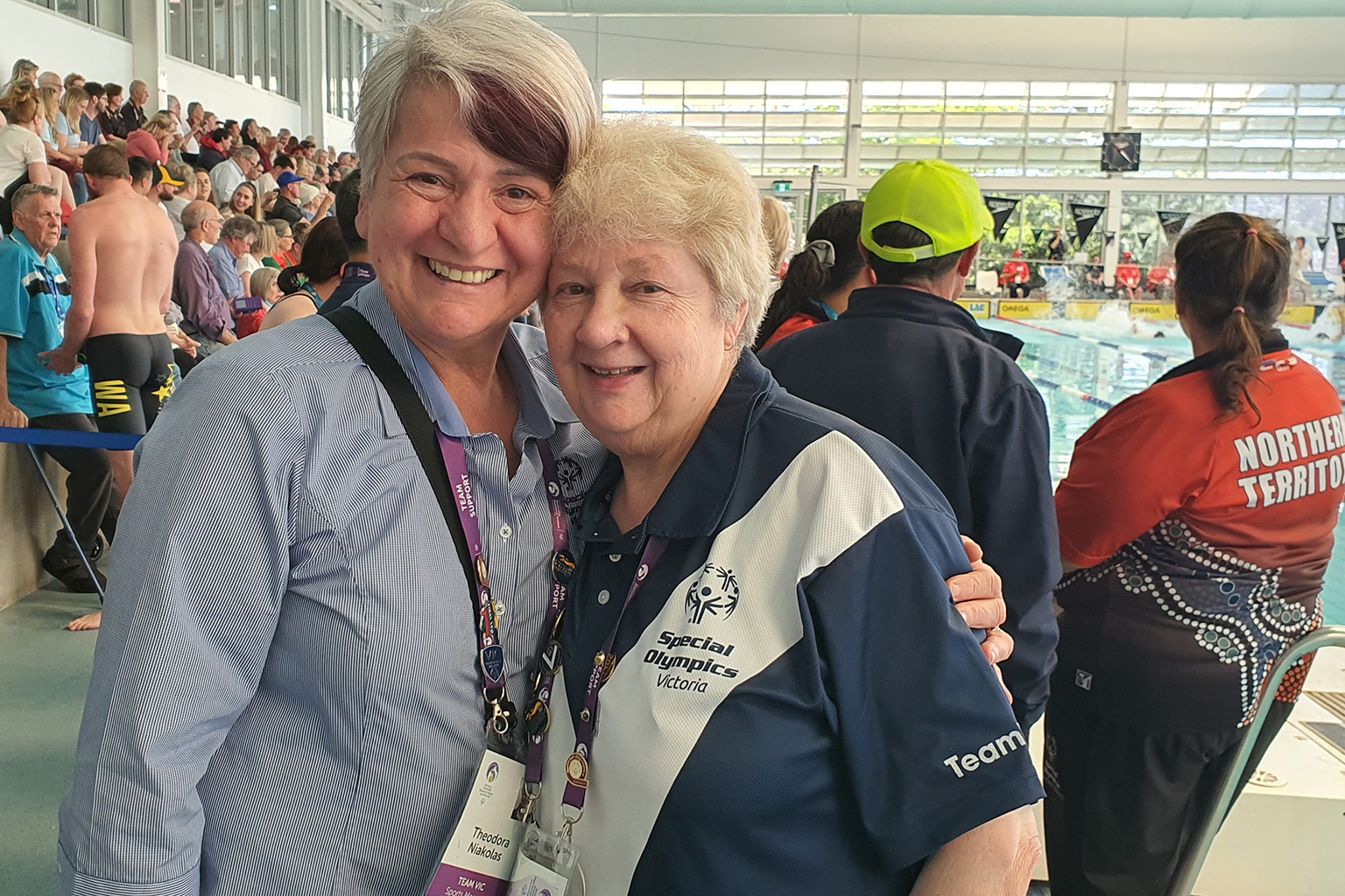 Two women stand together near an indoor swimming pool, with a large crowd in the stands behind them.