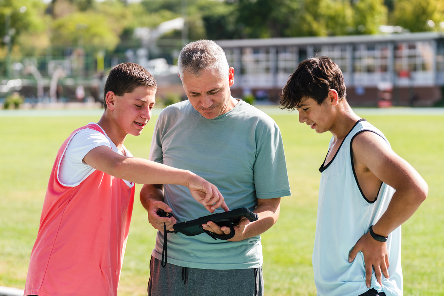 two teenage boys stand on an athletics track with an older male coach looking over an iPad.