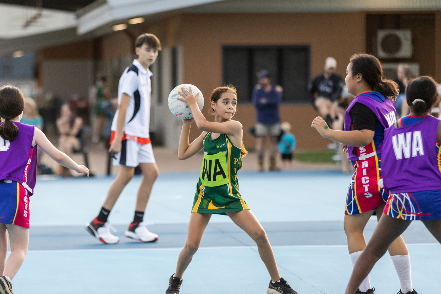 A young girl prepares to pass a netball while a netball umpire watches in the background