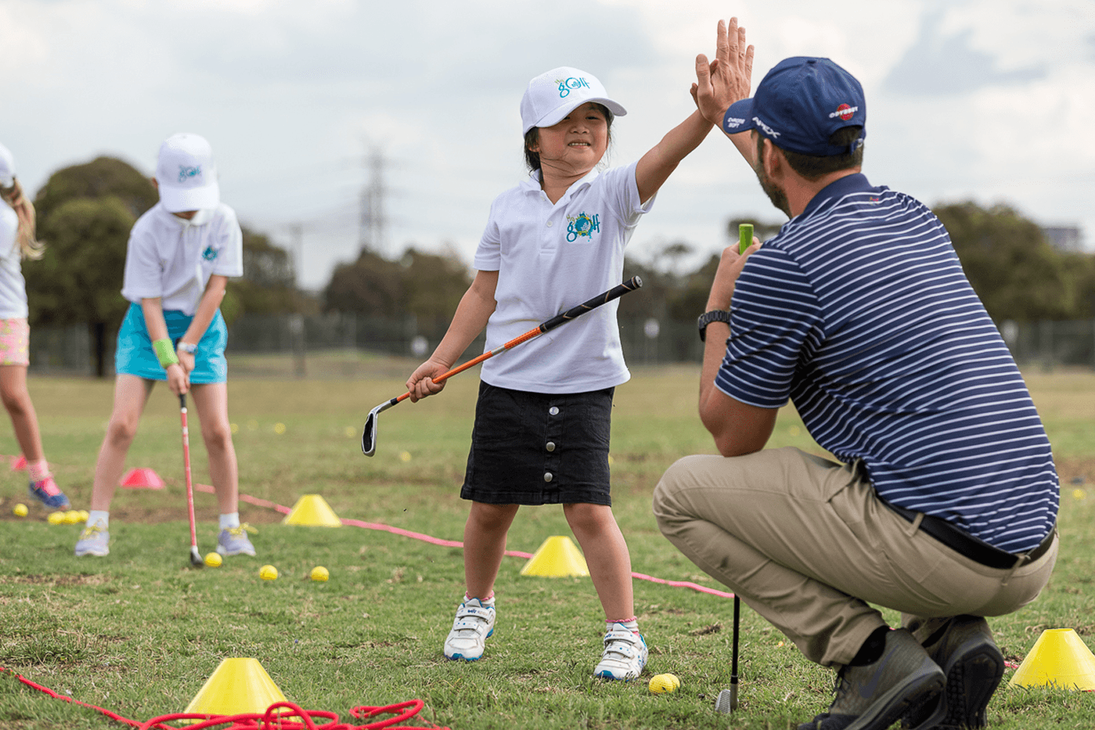 Young girl wearing white cap and golf shirt holds a golf club while high-fiving a golf coach.