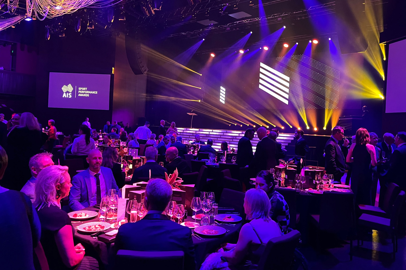 People seated at round tables in front of a large, boldly lit stage.