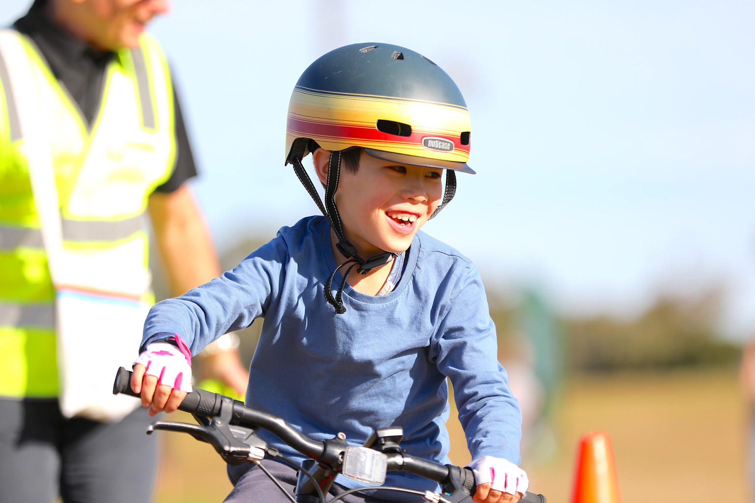 A young boy wearing a bicycle helmet while riding a bike. A man wearing a bright safety vest in the background.