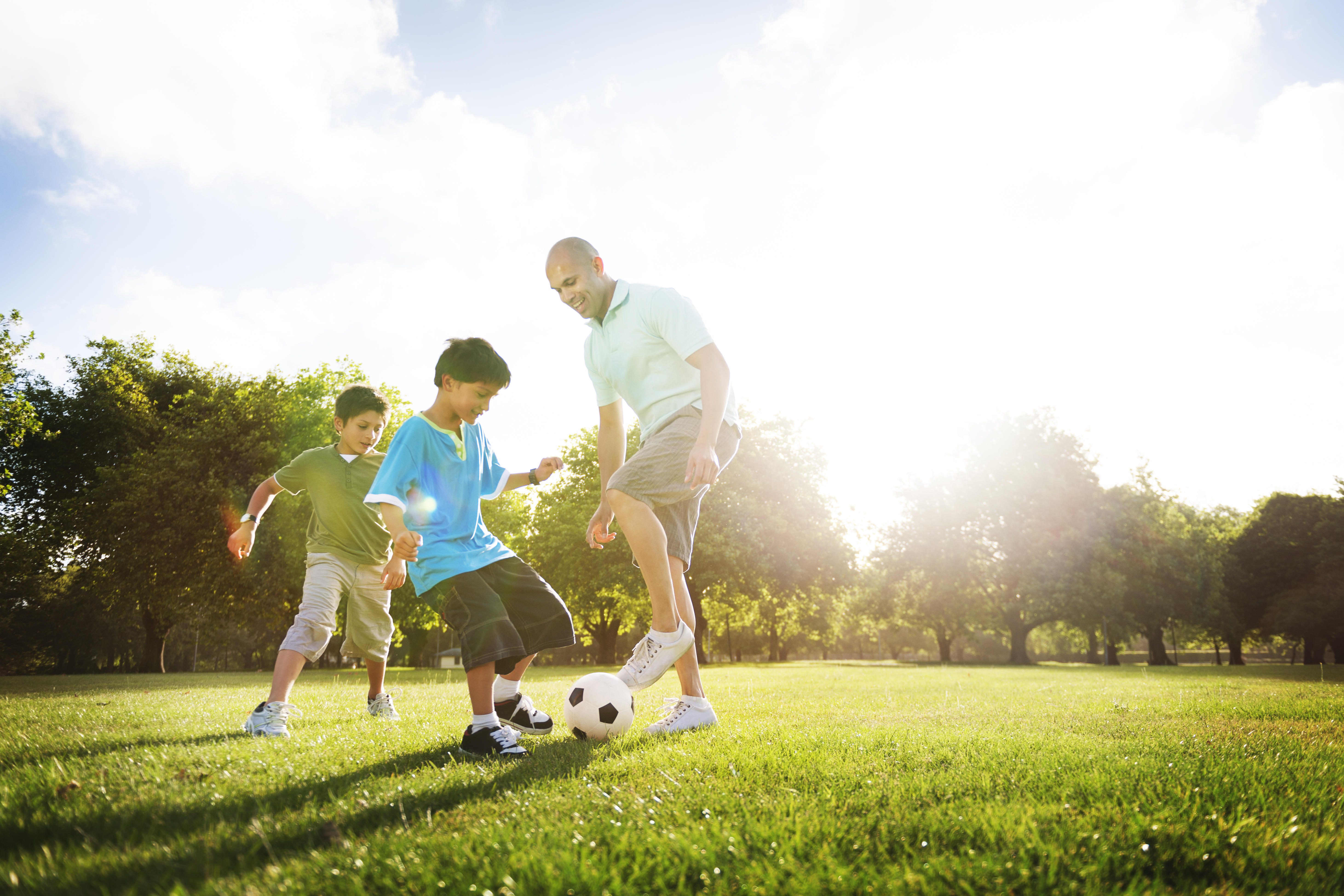 A father and two children kicking a soccer ball around a park