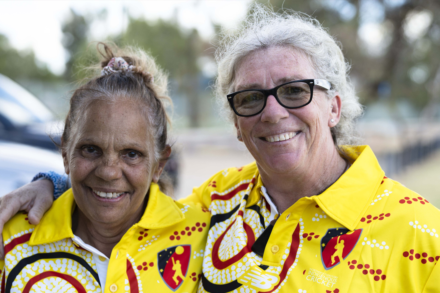 Two women in bright yellow, red and black indigenous-themed cricket shirts