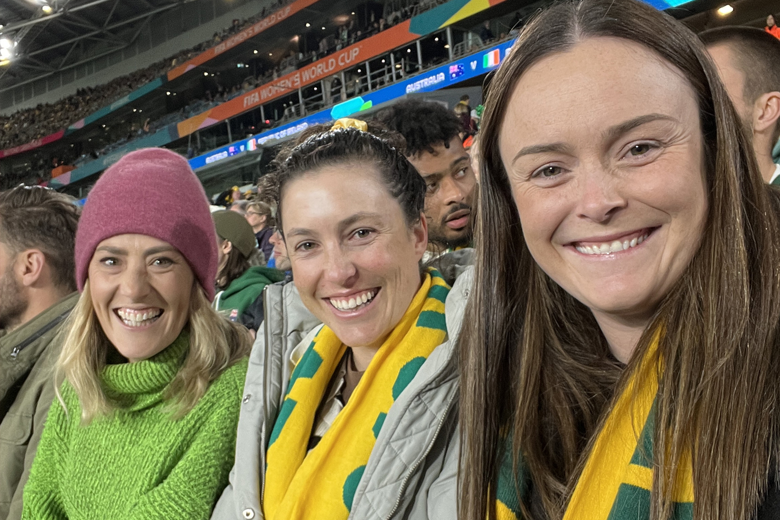 Three women sitting together wearing Matildas scarves at Stadium Australia.
