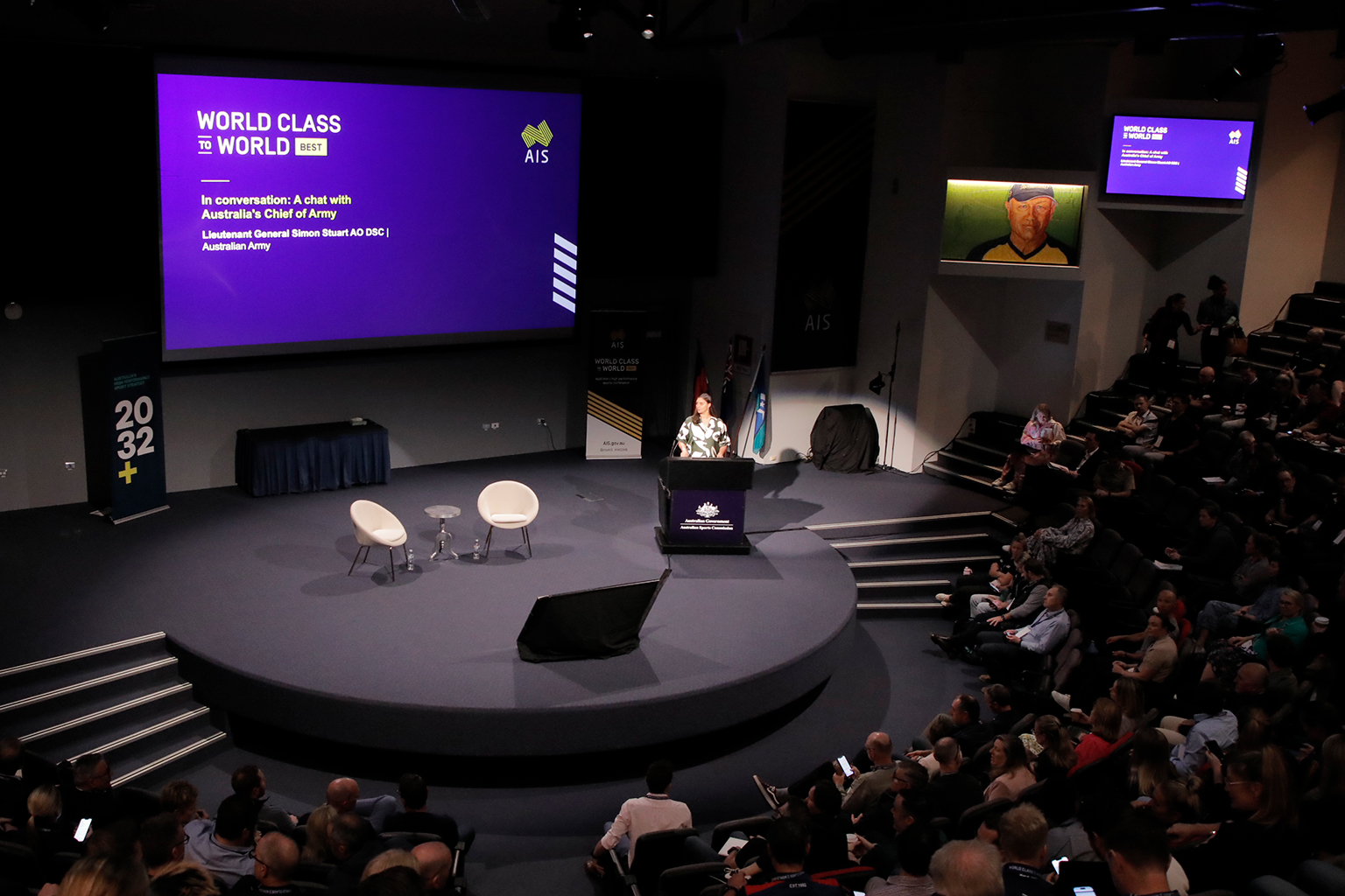 View across a full auditorium, showing a woman behind the lectern on stage.