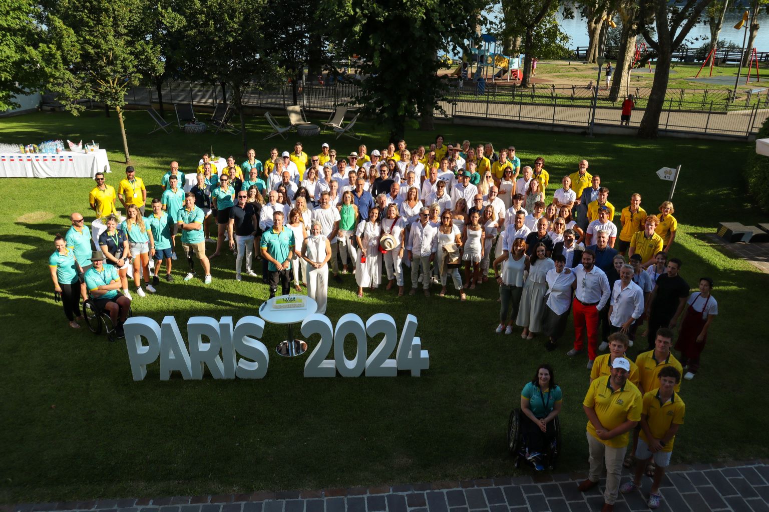 A group of people in white, green and gold, stand together behind standing letters that read 'PARIS 2024'