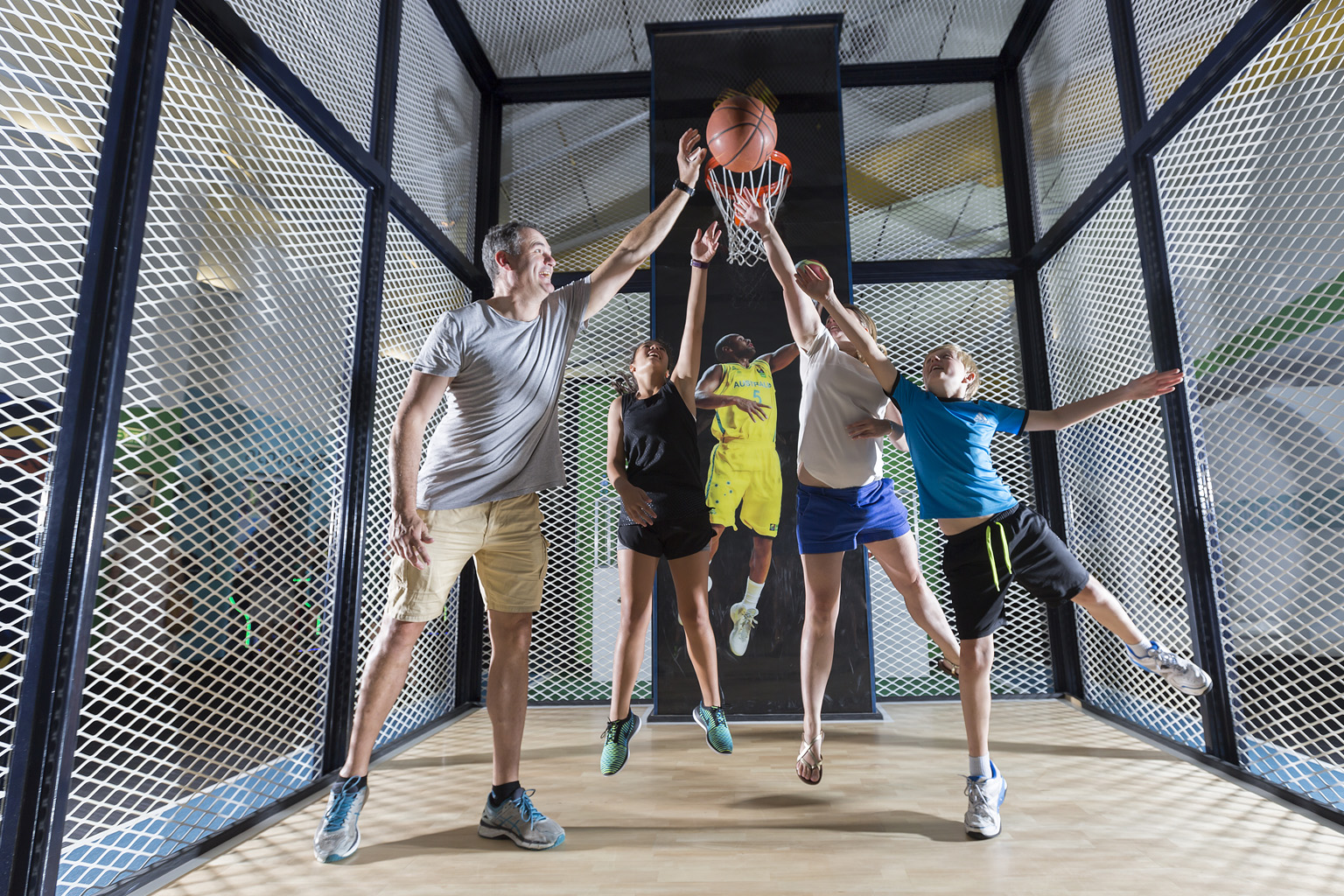 An adult and three children leap for a basketball in front of a basketball ring, inside a caged enclosure at Sportex