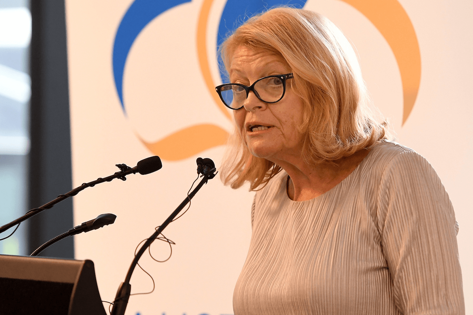 Lynne Anderson stands at a lectern speaking into microphones