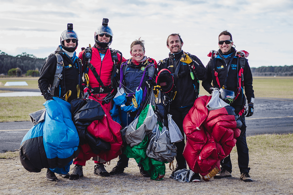 A group of five skydivers, with Jules McConnel centre, stand beside a tarmac with parachutes and skydiving equipment.