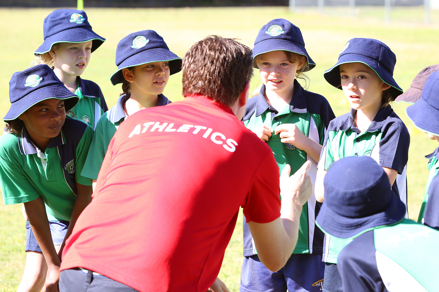 A man wearing a shirt with the word 'athletics' across the back speaks to school children.