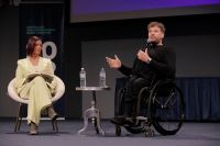 A woman seated next to a man in a wheelchair, speaking into a microphone
