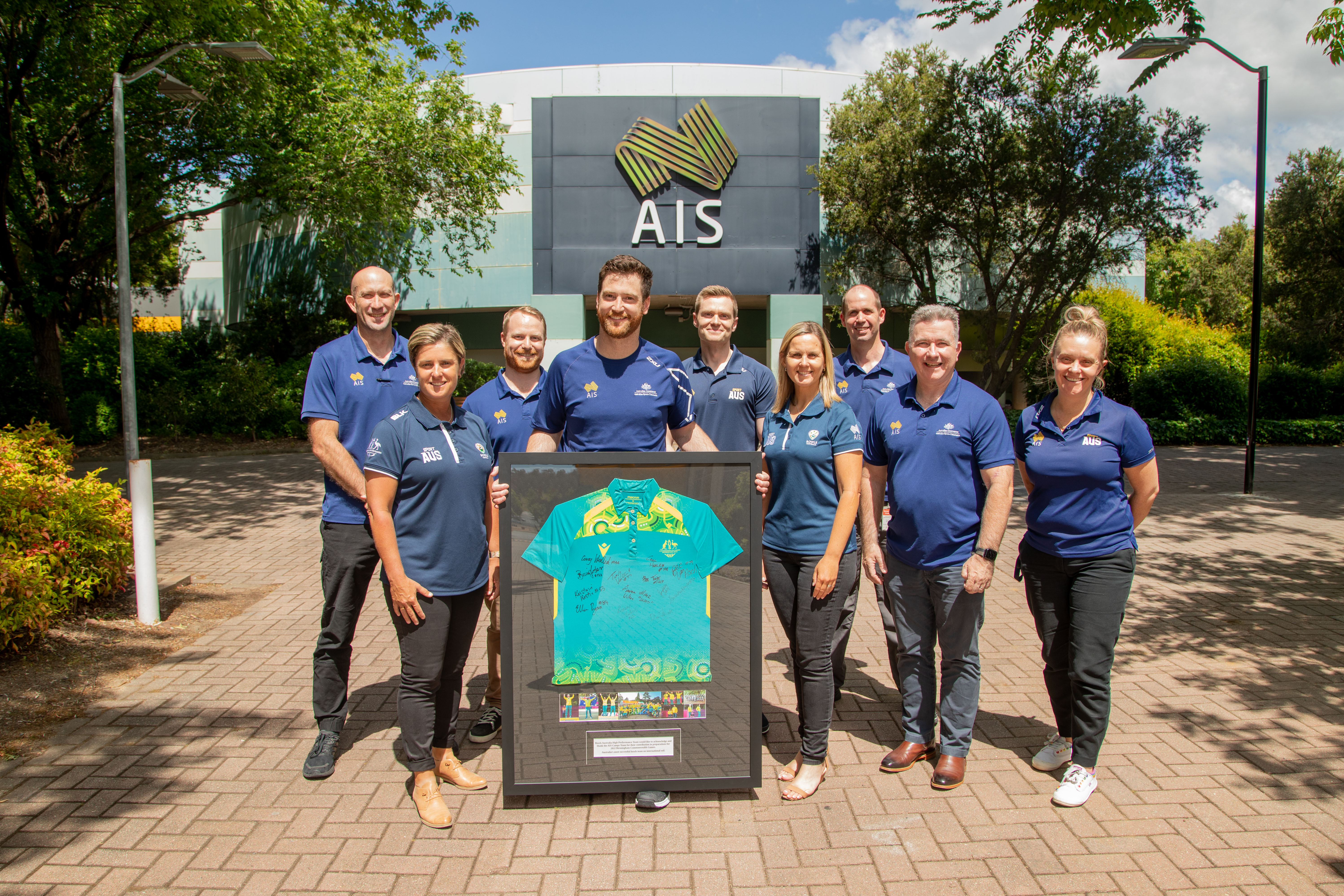 Image of people from the AIS and Bowls Australia wearing blue shirts stand in front of an AIS sign with a framed jersey