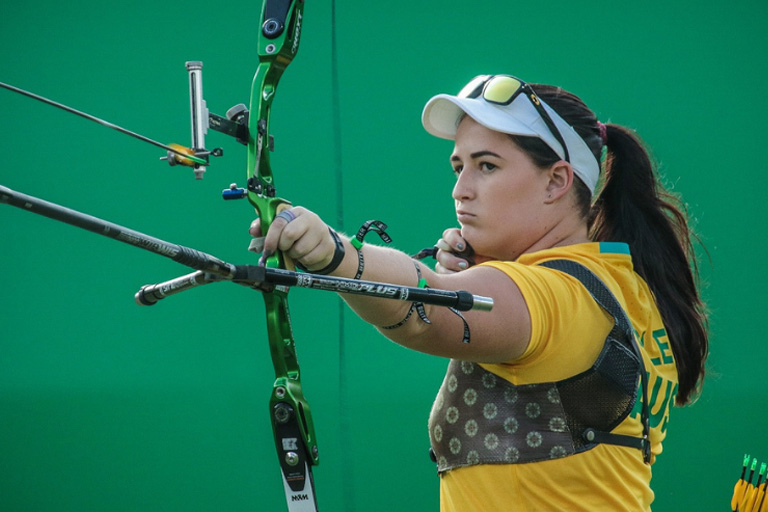 Woman archer standing in front of a green wall, wearing a yellow shirt and white hat, holding a bow