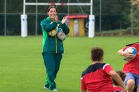 A woman claps for junior rugbyy players while holding two rugby balls to her body.