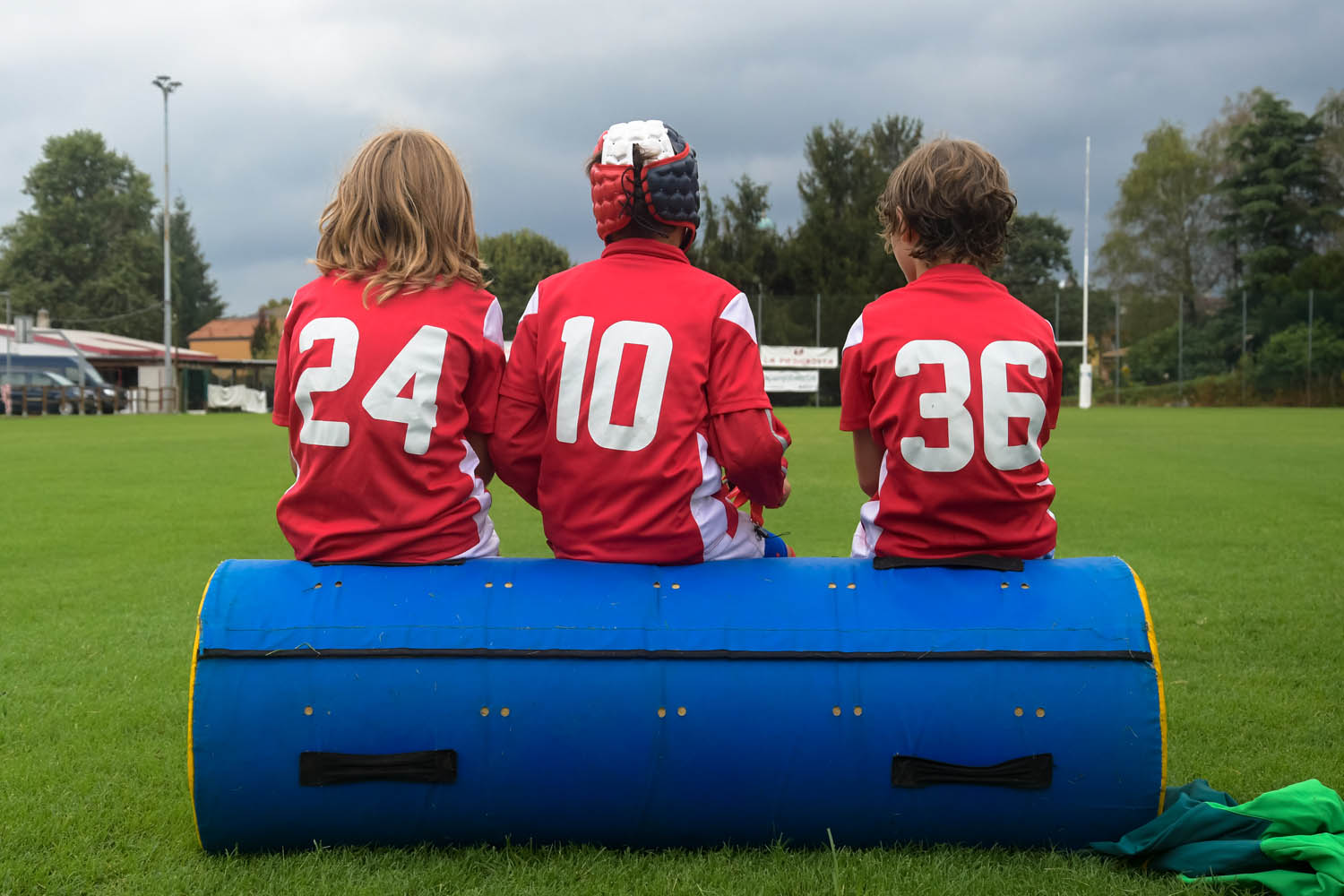 Three children wearing red rugby jerseys sit on post padding with their backs to the camera 