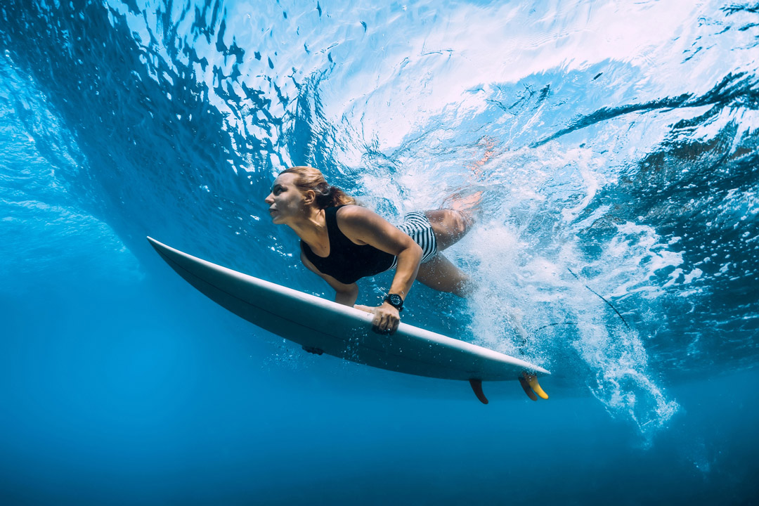 Underwater view of a woman swimming under a wave with a surfboard.